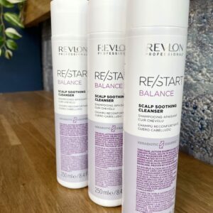 Three 250 ml bottles of Re/Start Balance Scalp Soothing Cleanser are displayed on a wooden surface, set against a textured wall background.
