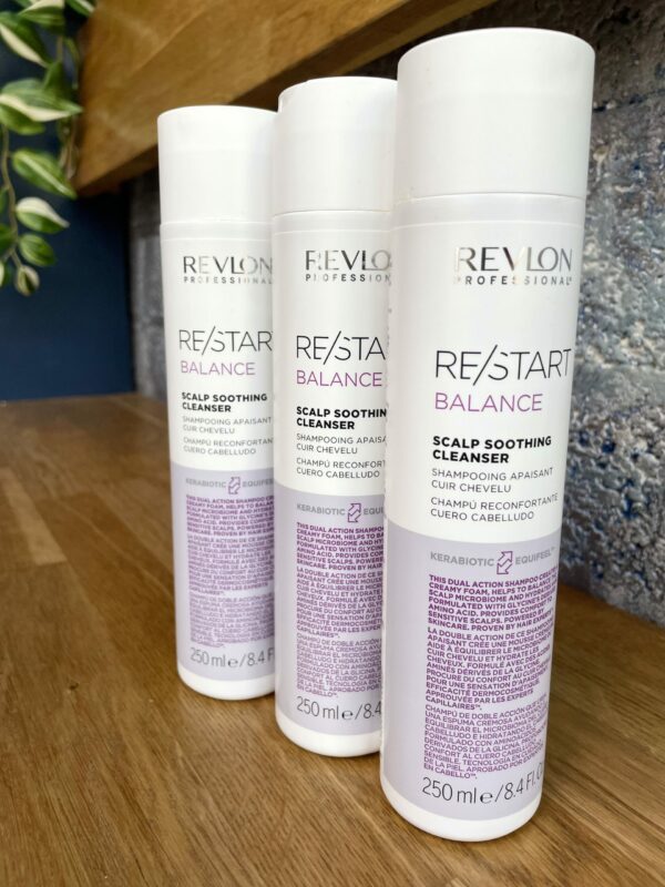 Three 250 ml bottles of Re/Start Balance Scalp Soothing Cleanser are displayed on a wooden surface, set against a textured wall background. Three 250 ml bottles of Re/Start Balance Scalp Soothing Cleanser are displayed on a wooden surface, set against a textured wall background.