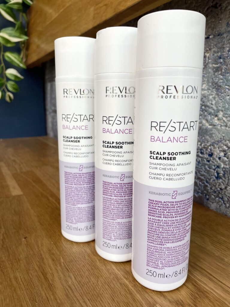 Three 250 ml bottles of Re/Start Balance Scalp Soothing Cleanser are displayed on a wooden surface, set against a textured wall background.
