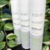 Three bottles of Re/Start Balance Purifying Micellar Shampoo are standing upright on a black surface, with green leafy plants in the background.