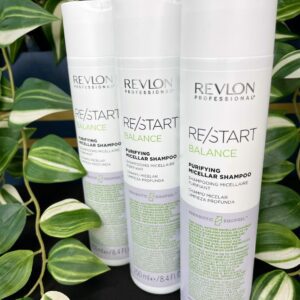 Three bottles of Re/Start Balance Purifying Micellar Shampoo are standing upright on a black surface, with green leafy plants in the background.