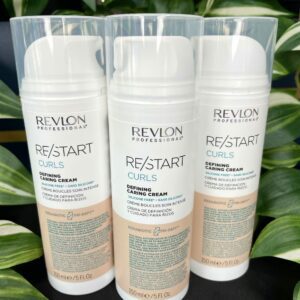 Three bottles of Re/Start Curls Defining Caring Cream (150ml) stand upright amid green leaves, emphasizing the nourishing benefits of this curl-defining cream.