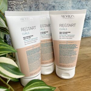 Three tubes of Re/Start Curls Multipurpose Gel-to-Oil (150 ml each) stand upright on a wooden surface beside green houseplants, ideal for defining and nourishing curls.