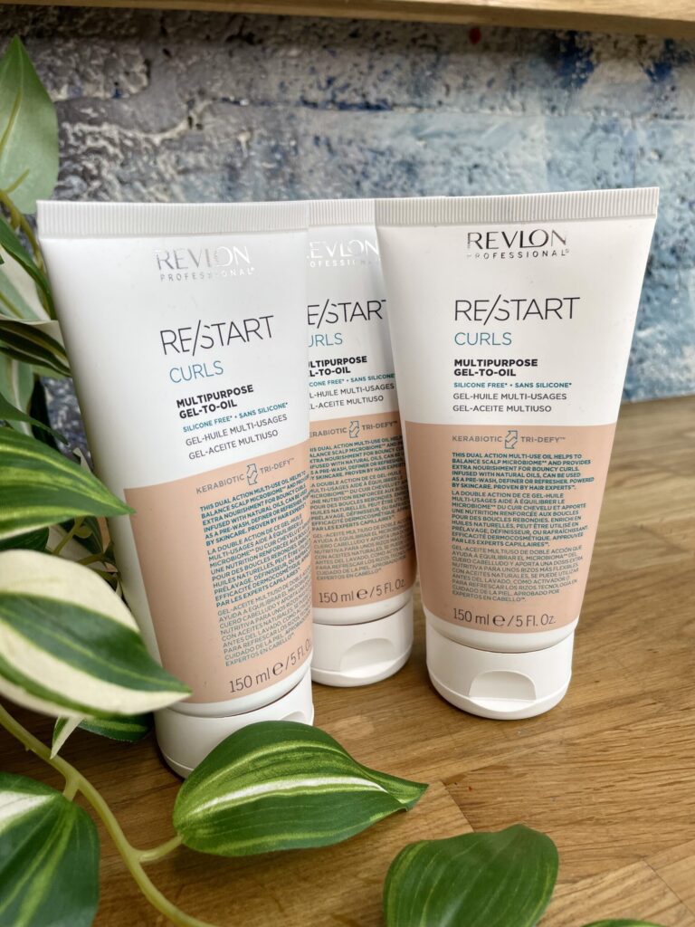 Three tubes of Re/Start Curls Multipurpose Gel-to-Oil (150 ml each) stand upright on a wooden surface beside green houseplants, ideal for defining and nourishing curls.