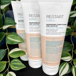 Three 200 ml tubes of Re/Start Curls Nourishing Conditioner and Leave-In stand upright with green plants in the background, emphasizing their nourishing benefits for curls.