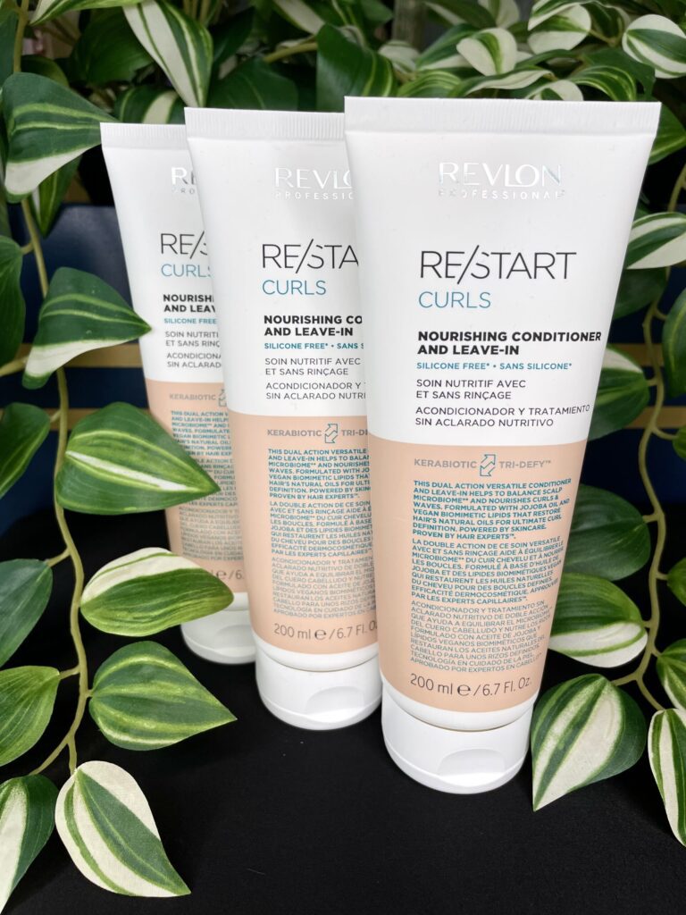 Three 200 ml tubes of Re/Start Curls Nourishing Conditioner and Leave-In stand upright with green plants in the background, emphasizing their nourishing benefits for curls.