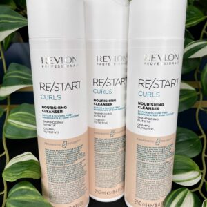 Three bottles of Re/Start Curls Nourishing Cleanser, 250 ml each, are displayed upright in front of lush green leafy plants.