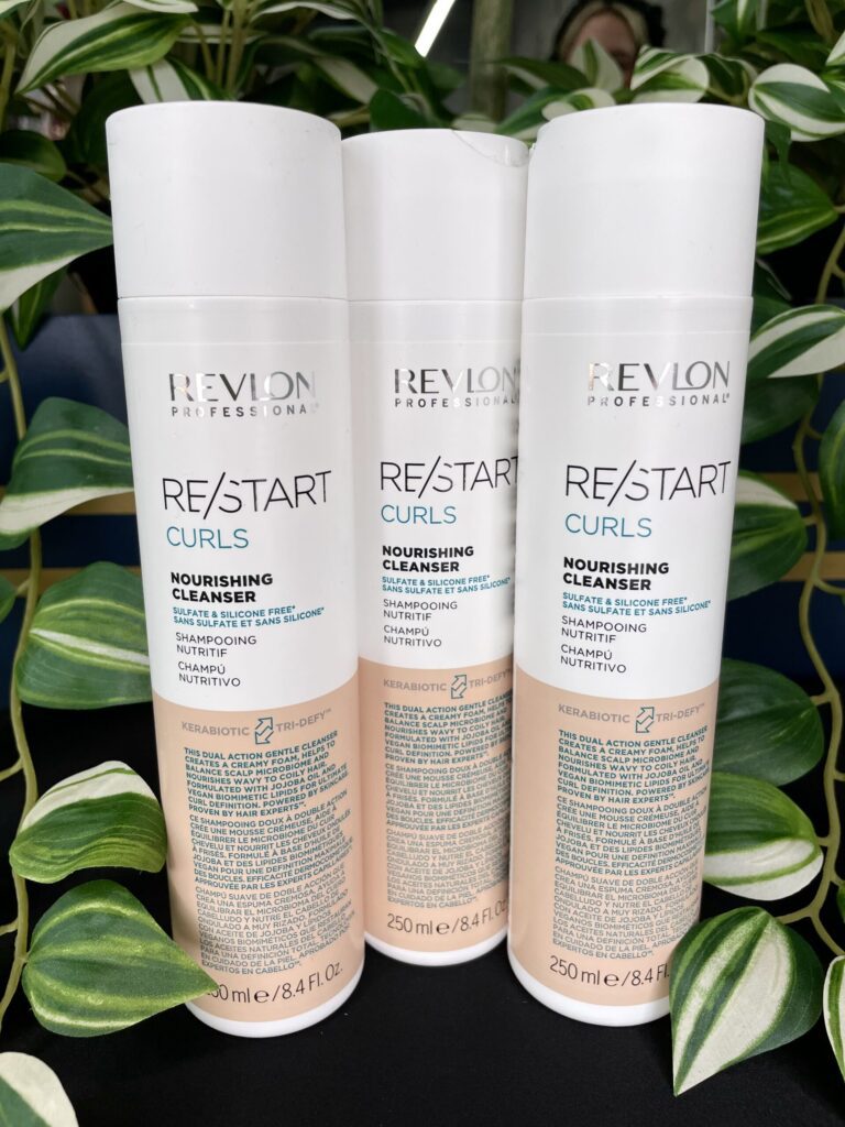 Three bottles of Re/Start Curls Nourishing Cleanser, 250 ml each, are displayed upright in front of lush green leafy plants.