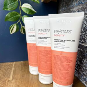 Three bottles of Re/Start Density Fortifying Weightless Conditioner are neatly arranged on a wooden surface beside a plant, with a blue wall in the background, highlighting these lightweight conditioners.