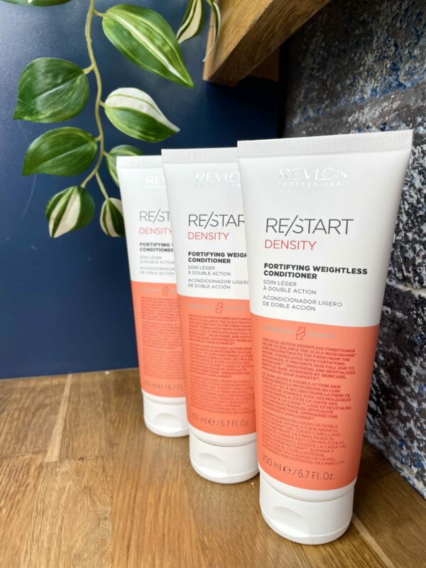 Three bottles of Re/Start Density Fortifying Weightless Conditioner are neatly arranged on a wooden surface beside a plant, with a blue wall in the background, highlighting these lightweight conditioners. Three bottles of Re/Start Density Fortifying Weightless Conditioner are neatly arranged on a wooden surface beside a plant, with a blue wall in the background, highlighting these lightweight conditioners.