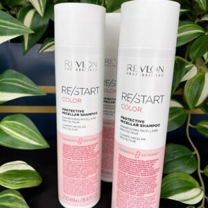 Three bottles of Re/Start Color Protective Micellar Shampoo are shown on a black surface with green and white leafy plants in the background, emphasizing the gentle care provided by this shampoo.