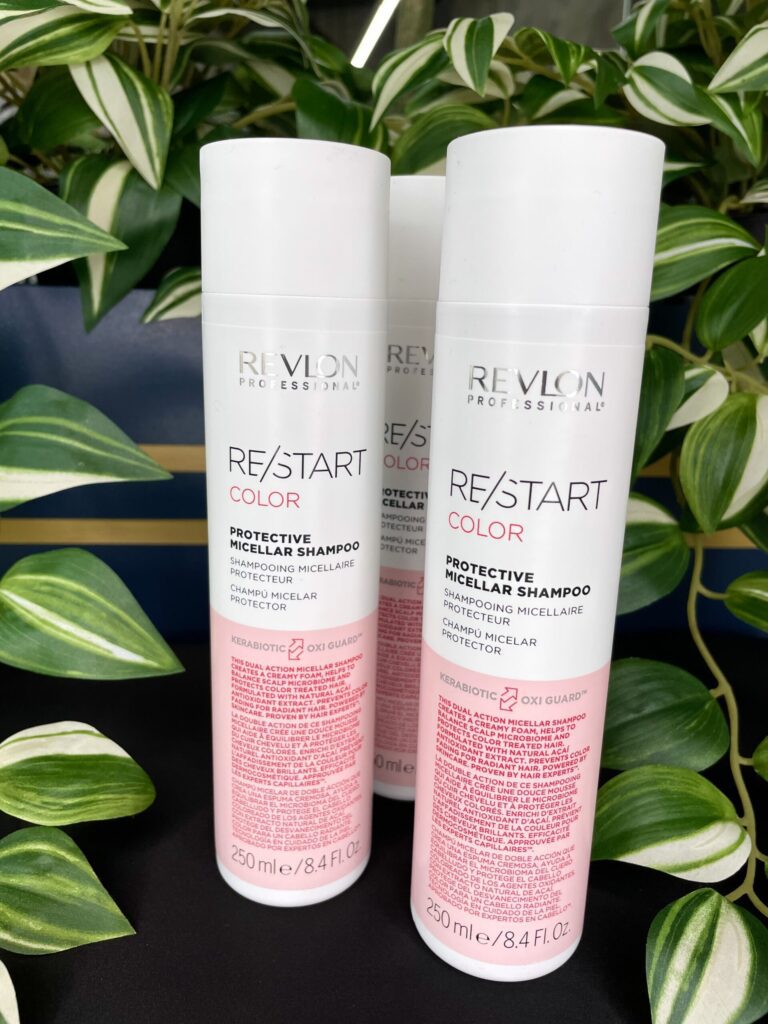 Three bottles of Re/Start Color Protective Micellar Shampoo are shown on a black surface with green and white leafy plants in the background, emphasizing the gentle care provided by this shampoo.