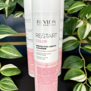 A 250 ml bottle of Re/Start Color Protective Gentle Cleanser is shown in front of green and white variegated plants, ideal for keeping hair vibrant and maintaining color.