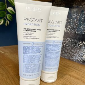 Two 200ml tubes of Re/Start Hydration Moisture Melting Conditioner are displayed on a wooden surface with a plant and blue wall behind, showcasing luxurious hydration and moisture care for your hair.