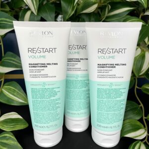 Three bottles of Re/Start Volume Magnifying Melting Conditioner stand upright on a black surface, with green plants visible in the background.