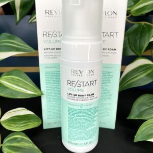A bottle of Re/Start Volume Lift-Up Body Foam, 165 ml, is displayed in front of its box with green leafy plants in the background.
