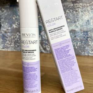 A bottle and box of Re/Start Color Anti-Brassiness Purple Drops are shown on a wooden surface, emphasizing its advanced formula for expert color care.