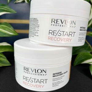 Two stacked tubs of Re/Start Recovery Intense Recovery Mask are displayed on a black surface, with lush green plants in the background.