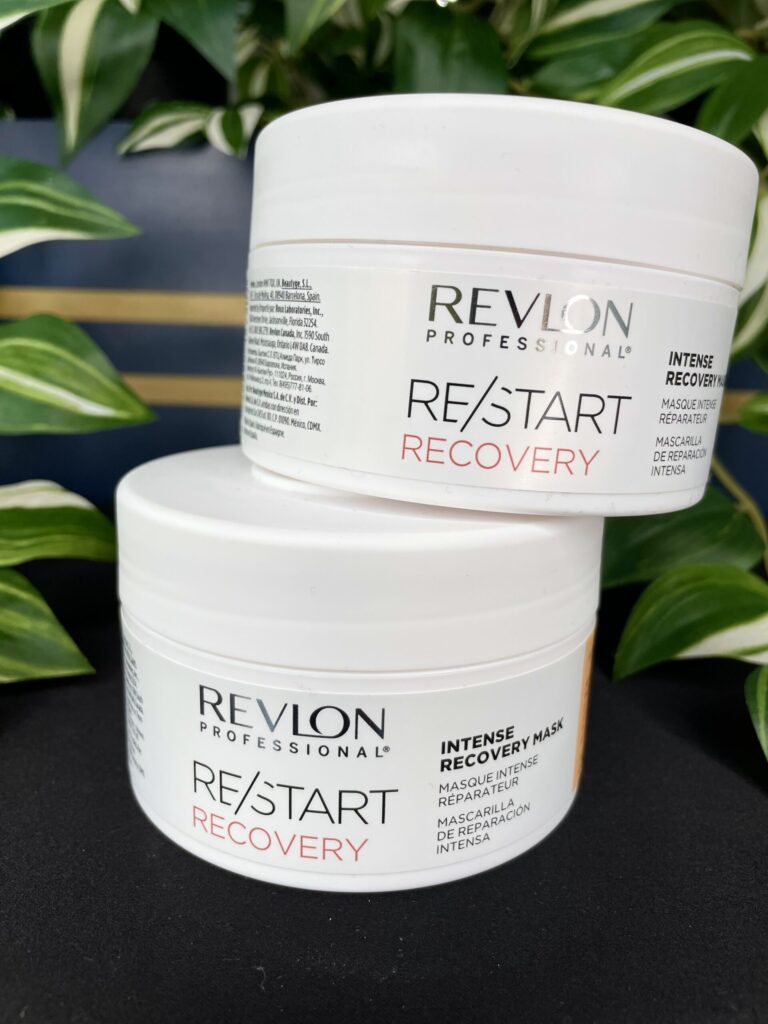 Two stacked tubs of Re/Start Recovery Intense Recovery Mask are displayed on a black surface, with lush green plants in the background.