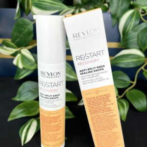 A bottle and box of Re/Start Recovery Anti-Split Ends Sealing Drops, 50ml, sit on a dark surface with green leafy plants in the background, emphasizing its anti-split ends recovery benefits.
