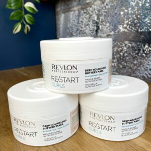 Three containers of Re/Start Curls Deep Nourishing Buttery Mask are stacked on a wooden surface, with a plant and blue wall in the background.