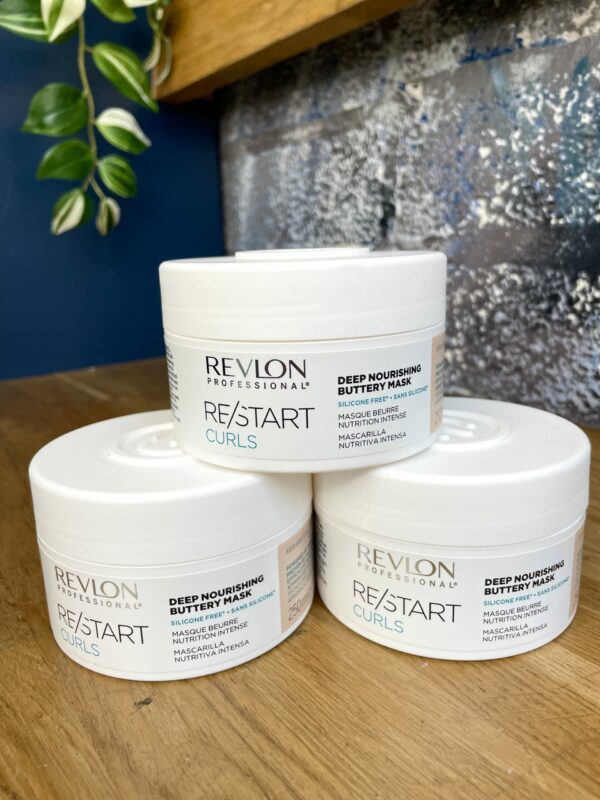 Three containers of Re/Start Curls Deep Nourishing Buttery Mask are stacked on a wooden surface, with a plant and blue wall in the background. Three containers of Re/Start Curls Deep Nourishing Buttery Mask are stacked on a wooden surface, with a plant and blue wall in the background.