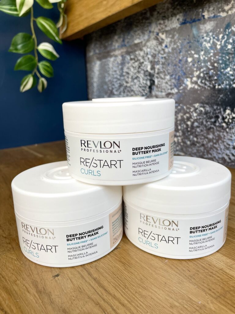 Three containers of Re/Start Curls Deep Nourishing Buttery Mask are stacked on a wooden surface, with a plant and blue wall in the background.