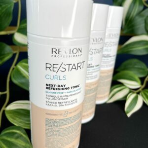 A bottle of Re/Start Curls Next-Day Refreshing Tonic is in focus, showcasing its curl-refreshing power, with two more bottles and green plants blurred in the background.