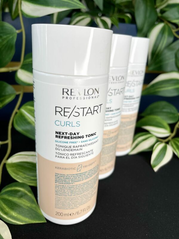 A bottle of Re/Start Curls Next-Day Refreshing Tonic is in focus, showcasing its curl-refreshing power, with two more bottles and green plants blurred in the background. A bottle of Re/Start Curls Next-Day Refreshing Tonic is in focus, showcasing its curl-refreshing power, with two more bottles and green plants blurred in the background.