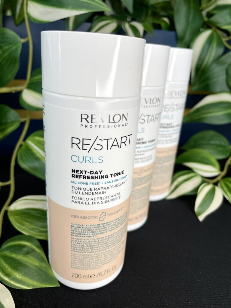 A bottle of Re/Start Curls Next-Day Refreshing Tonic is in focus, showcasing its curl-refreshing power, with two more bottles and green plants blurred in the background.