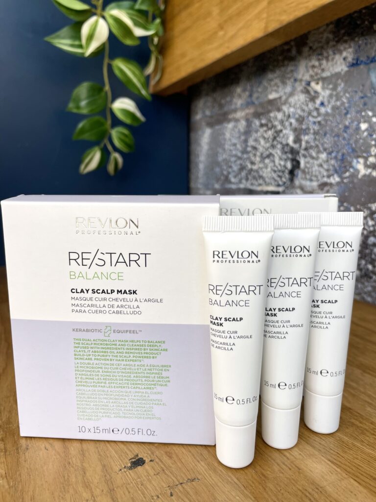 Three tubes and a box of Re/Start Balance Clay Scalp Mask sit on a wooden surface with green leaves in the background, highlighting this mask designed to help balance scalp health.