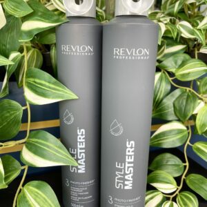 Two tall gray bottles of Style Masters Photo Finisher Hairspray 500ml are set amidst lush green foliage.