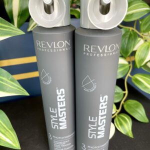 Two grey cans of Style Masters Photo Finisher Hairspray 200ml stand upright on a black surface, with green leafy plants in the background.