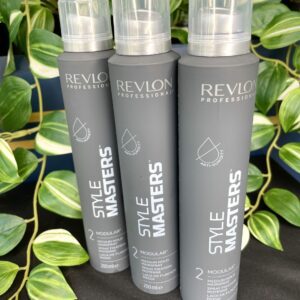 Three cans of Style Masters Modular Hairspray 200ml stand upright before a backdrop of lush green leafy plants.