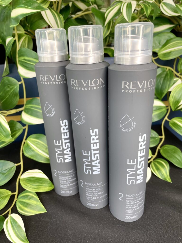 Three cans of Style Masters Modular Hairspray 200ml stand upright before a backdrop of lush green leafy plants.