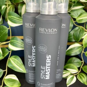 Three bottles of Style Masters Glamourama Shine Spray 300ml stand upright among green and white leafy plants.