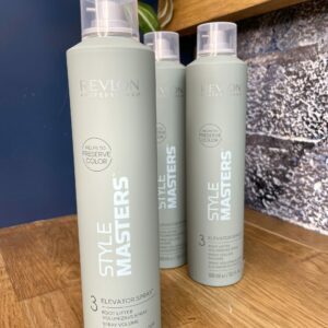 Three bottles of Style Masters Elevator Spray 300ml are displayed on a wooden shelf, with a plant and a blue textured wall in the background.