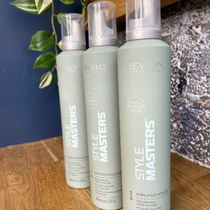 Three bottles of Style Masters Amplifier Mousse 300ml sit on a wooden shelf, with a green leafy plant in the background.
