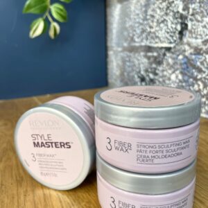 Three 85g containers of Style Masters Fiber Wax are stacked on a wooden surface, with one lying flat next to them. A green plant hangs in the background.