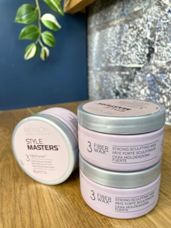 Three 85g containers of Style Masters Fiber Wax are stacked on a wooden surface, with one lying flat next to them. A green plant hangs in the background. Three 85g containers of Style Masters Fiber Wax are stacked on a wooden surface, with one lying flat next to them. A green plant hangs in the background.