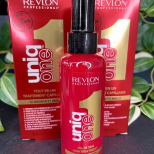 A red bottle of UniqOne All in One Hair Treatment Spray Original 150ml is shown in front of its packaging, with green leaves in the background.