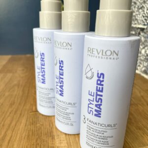 Three bottles of Style Masters Fanaticurls Curl Activator are lined up on a wooden surface, with a plant and a stone wall in the background.