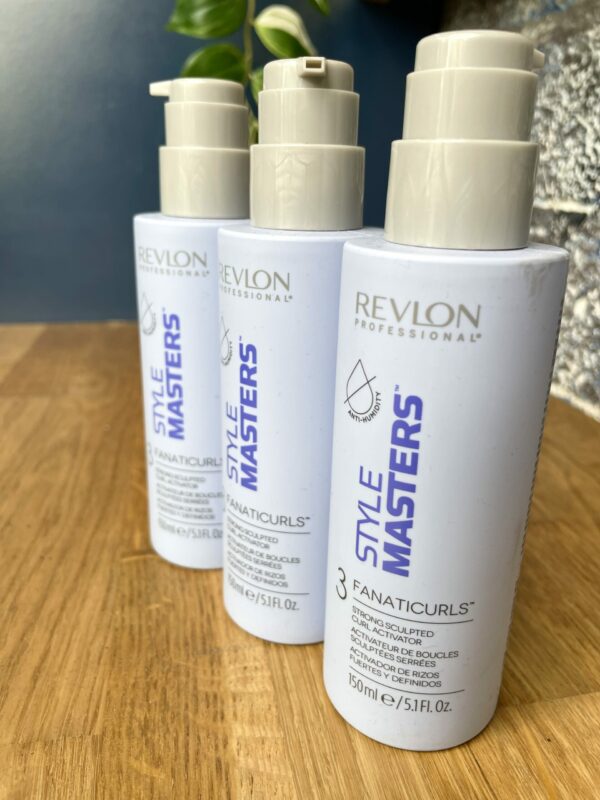Three bottles of Style Masters Fanaticurls Curl Activator are lined up on a wooden surface, with a plant and a stone wall in the background. Three bottles of Style Masters Fanaticurls Curl Activator are lined up on a wooden surface, with a plant and a stone wall in the background.