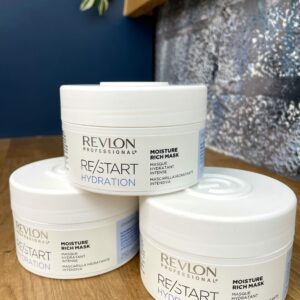 Three white containers of Re/Start Hydration Moisture Rich Mask are stacked on a wooden surface, set against a blue and metallic background.