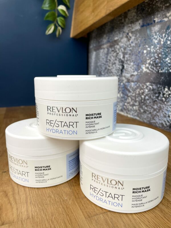 Three white containers of Re/Start Hydration Moisture Rich Mask are stacked on a wooden surface, set against a blue and metallic background.