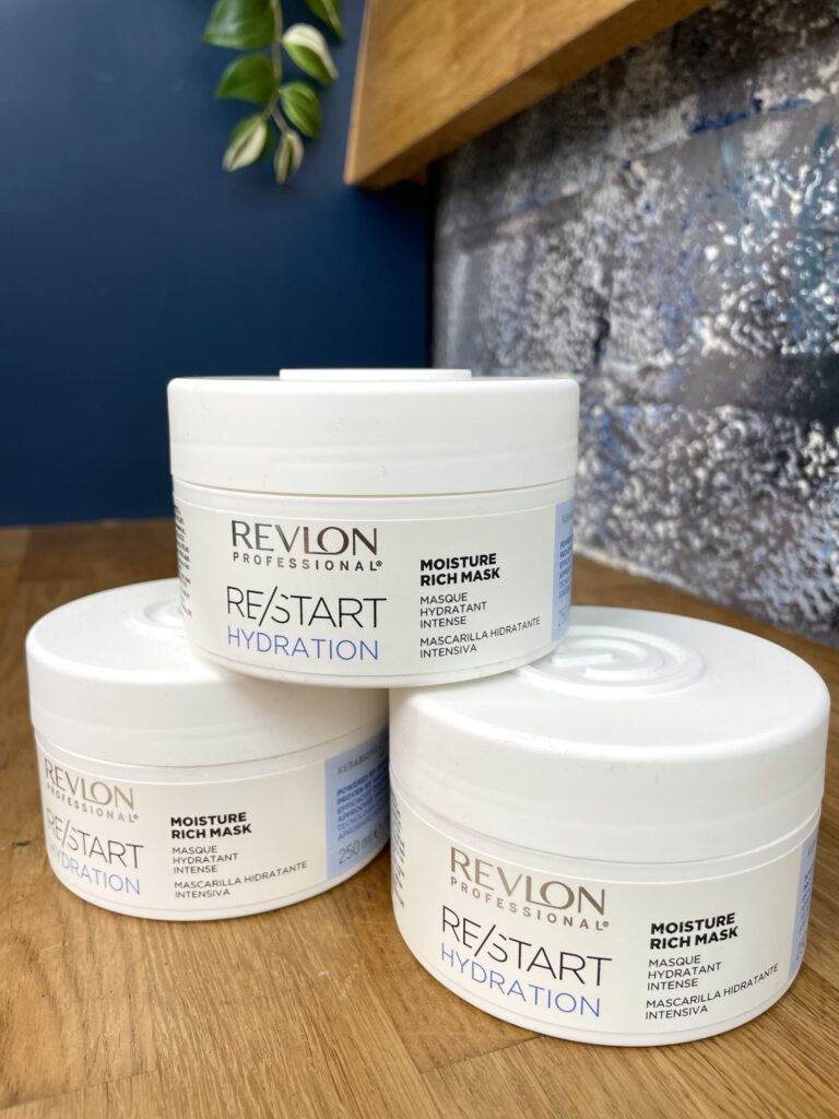 Three white containers of Re/Start Hydration Moisture Rich Mask are stacked on a wooden surface, set against a blue and metallic background.