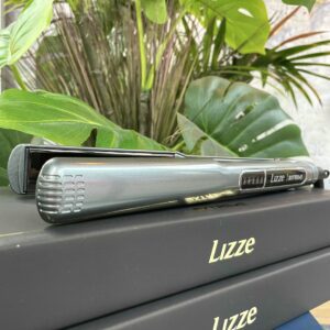 A Lizze Extreme Hair Straightener is placed on two stacked black boxes labeled Lizze Extreme Hair Straighteners, with green plants in the background.