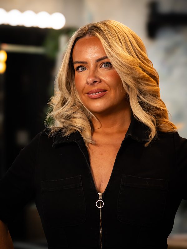A woman with blonde, wavy hair wearing a black zip-up top stands indoors, looking at the camera with a neutral expression.