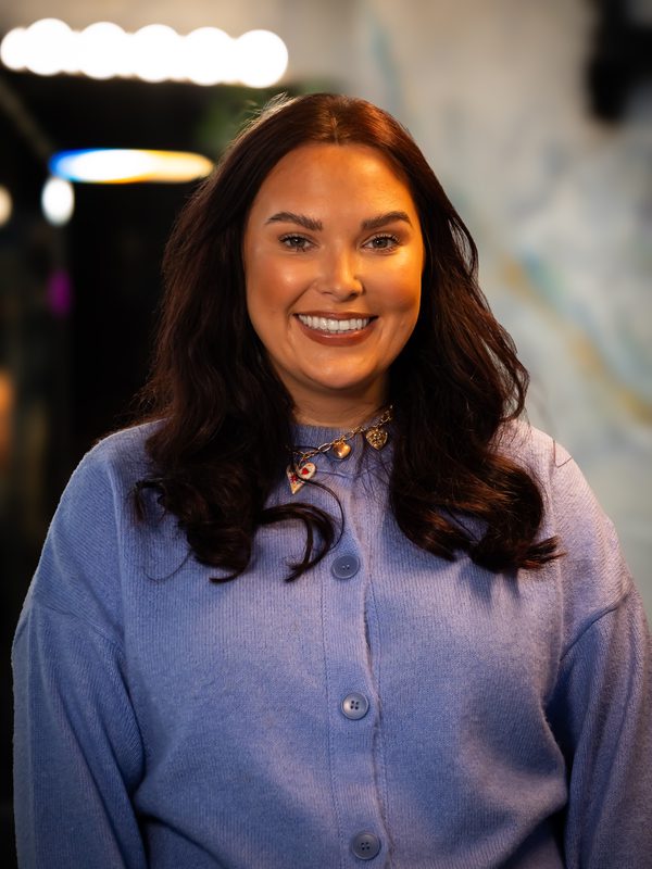 A woman with long brown hair wearing a light purple button-up cardigan and a necklace, standing indoors and smiling at the camera.