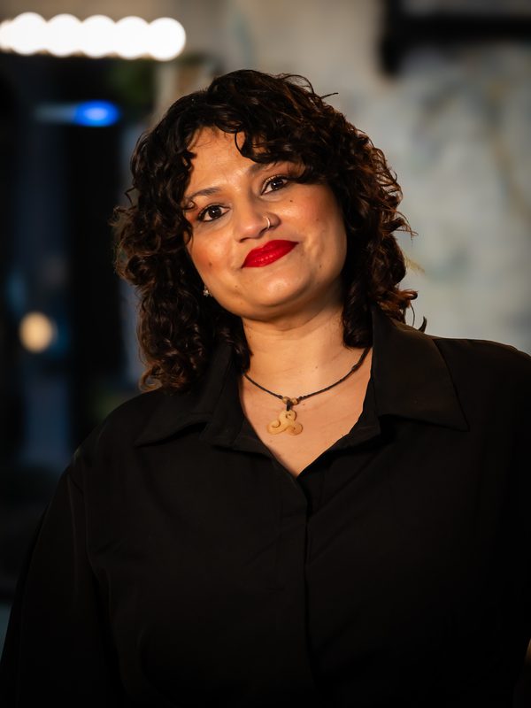 A woman with curly hair and red lipstick, wearing a black top and a necklace, stands indoors with a blurred background.